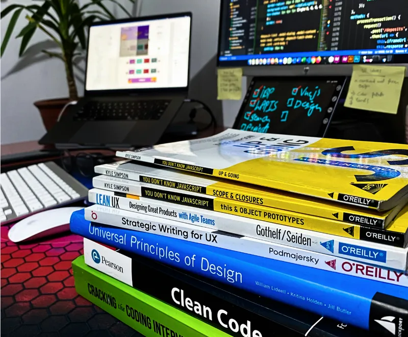 Stack of software development books in front of a laptop and monitors showing code.