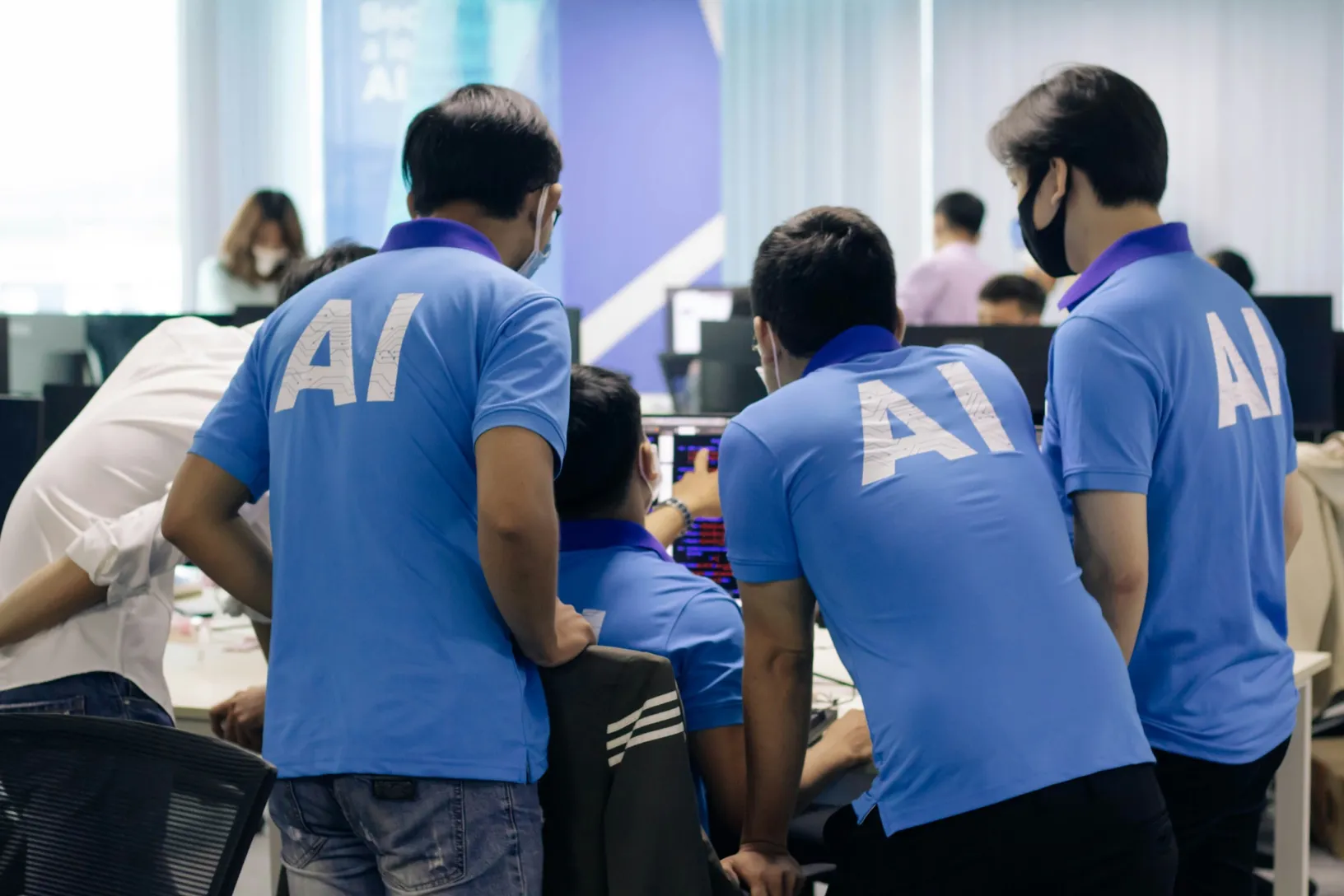 Team members in shirts printed with AI gathered around a colleague's computer.