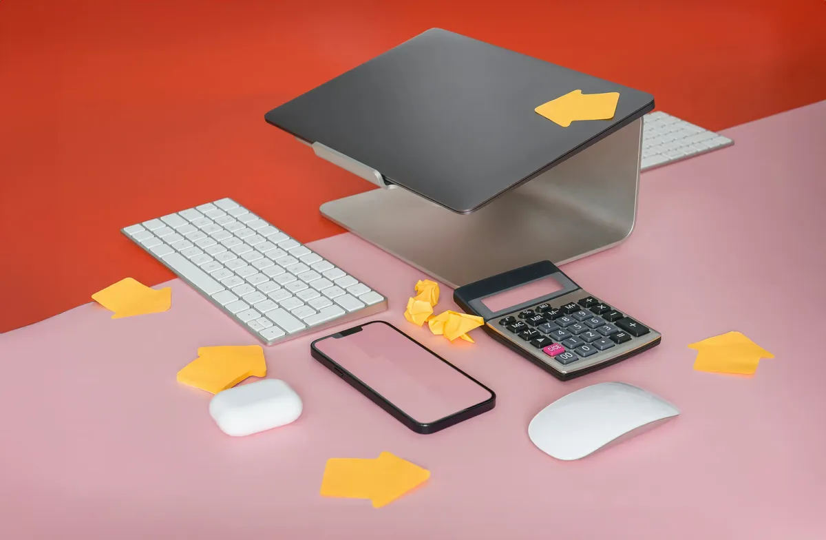 Modern workspace setup with wireless keyboard, smartphone, calculator, wireless mouse, AirPods case, and laptop stand on a pink and red desk with yellow arrow sticky notes.